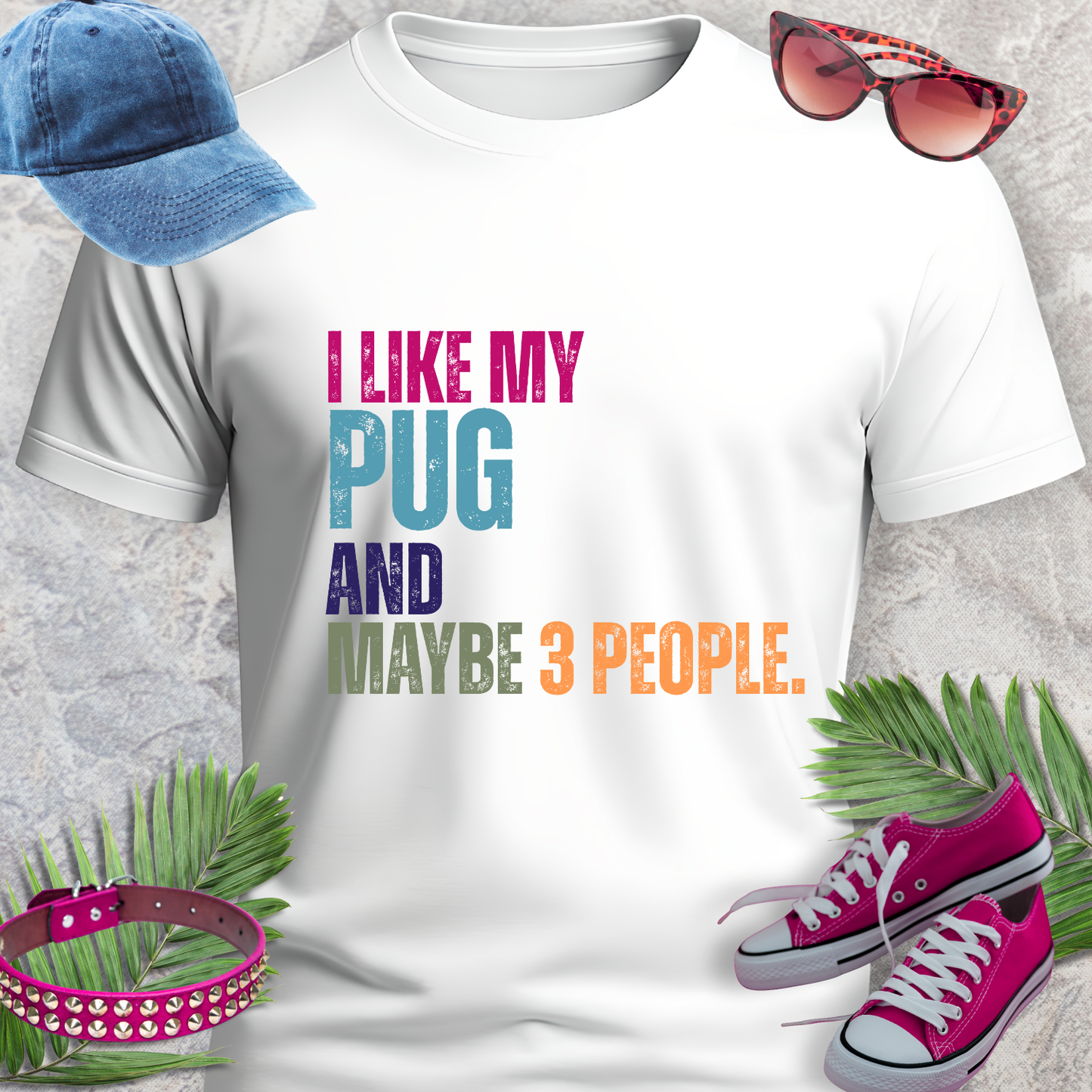 White t-shirt with colorful text 'I LIKE MY PUG AND MAYBE 3 PEOPLE.' displayed with accessories on a textured surface.