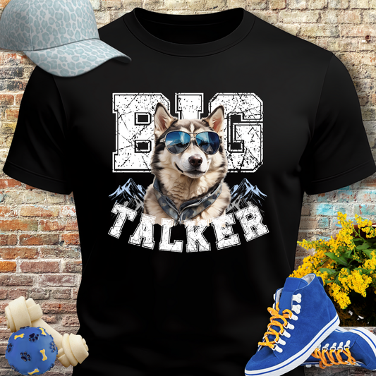 Black t-shirt with a dog graphic and 'BIG TALKER' text, placed against a brick wall with shoes and a cap on either side.