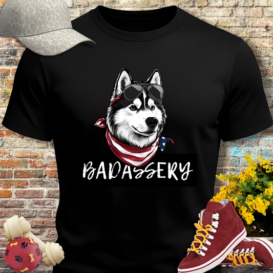 Black t-shirt with a dog graphic and 'BADASSERY' text, placed against a brick wall with shoes and a cap on either side.
