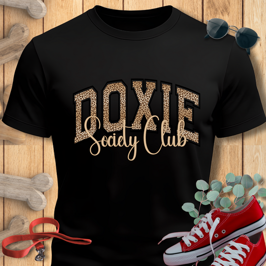 Black t-shirt with 'Doxie Society Club' text on a wooden surface with red sneakers and a dog leash.