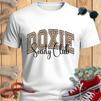 White t-shirt with 'Doxie Society Club' design on a wooden surface with red sneakers and sunglasses.