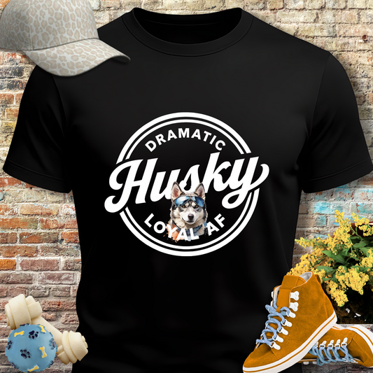 Black t-shirt with 'Dramatic Husky Local AF' design, placed against a brick wall with accessories.