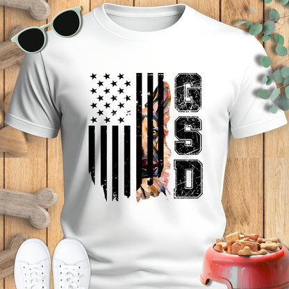 White t-shirt with American flag and 'GSD' design on a wooden surface with sunglasses and dog toys.