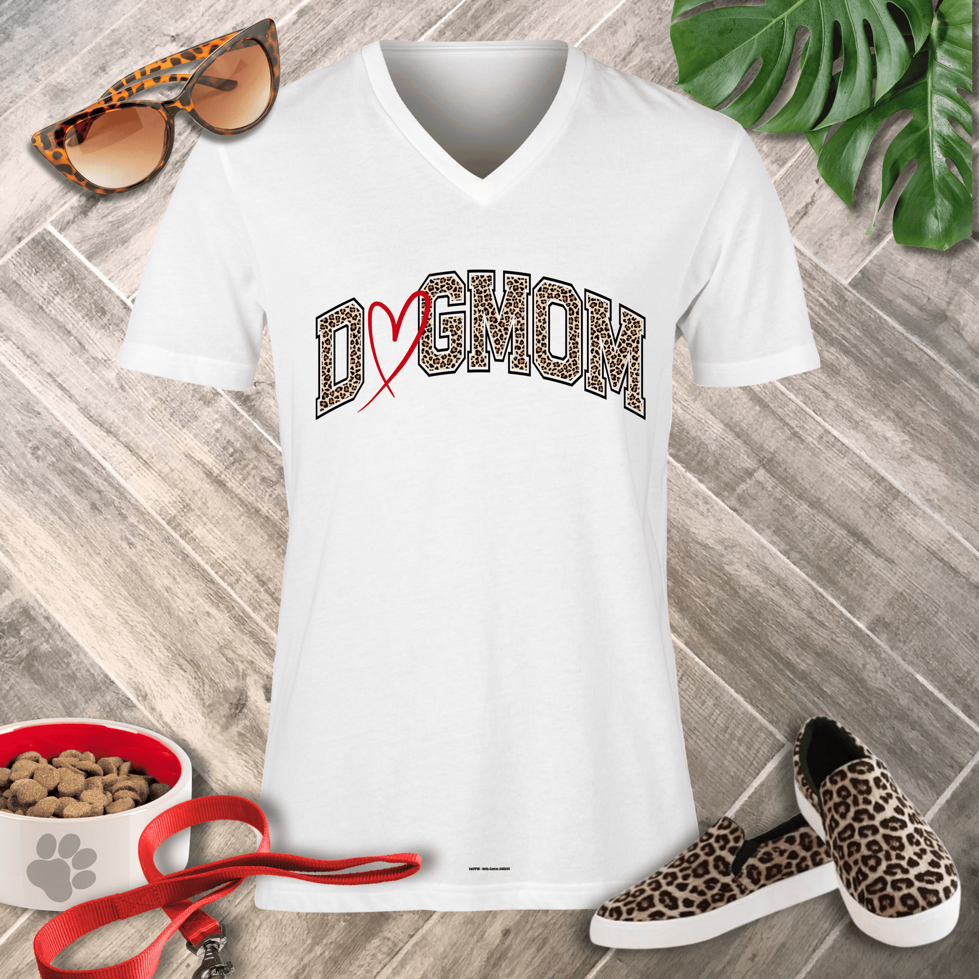 A white V-neck t-shirt with a 'DOG MOM' graphic in leopard print and a red heart, styled in a flat lay on a gray wood floor with leopard print sunglasses, shoes, a pet leash, a bowl of dog food, and a green plant.